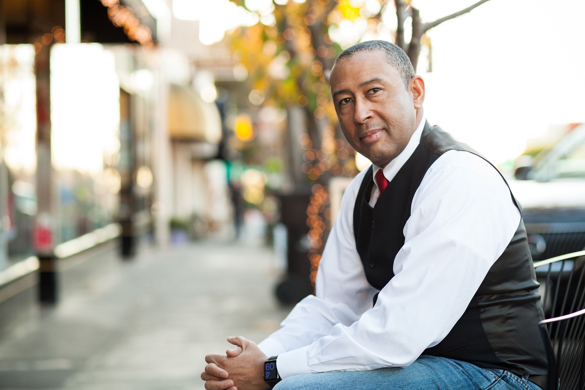 jay ward sitting in a cafe chair in downtown san francisco. he is wearing a white button-down shirt, red tie, black vest, and jeans.